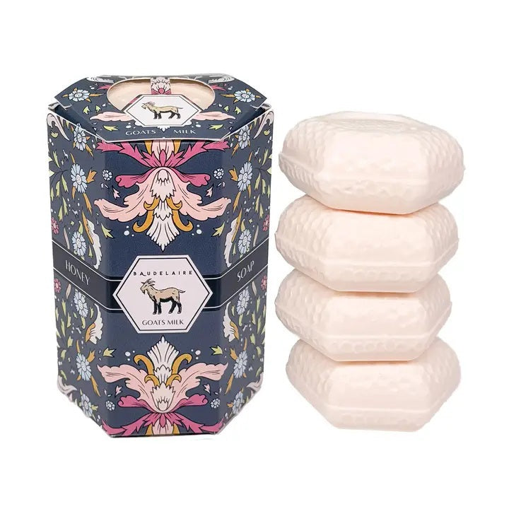 A decorative BAUDELAIRE - GOATS MILK HONEY SOAP 1.4 OZ QUARTET box sits beside a stack of four pale pink, hexagon-shaped soap bars. The floral-patterned box features an illustration of a goat, drawing attention to this gentle triple-milled soap from BAUDELAIRE, crafted with nourishing goat milk and natural honey.