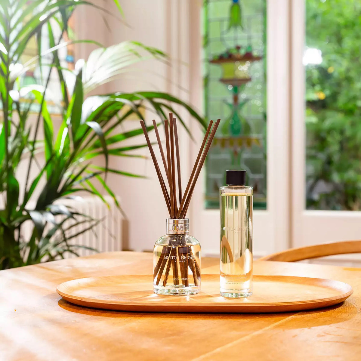 A clear glass bottle filled with yellow liquid, labeled &quot;MOLTON BROWN - ORANGE AND BERGAMOT DIFFUSER,&quot; features several long, thin, dark brown diffuser reeds extending from its neck. The reed diffuser boasts a hint of Sevillian orange and neroli. A metallic ring at its opening gives it an elegant and classy appearance.