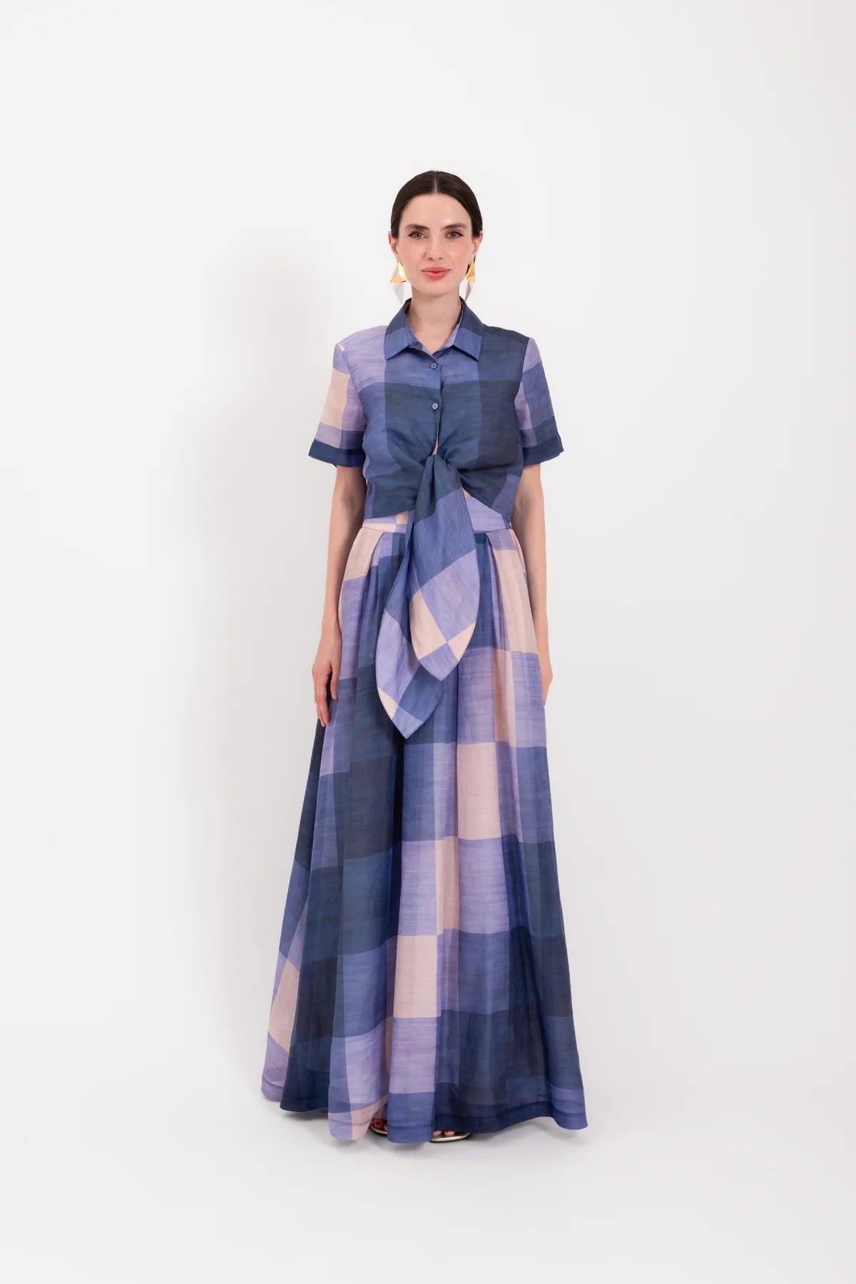 A woman wearing the LAVI - FRESIA Tie Front Cropped Top in Quadri, featuring a blue and light purple plaid pattern with short sleeves, a buttoned front, and an oversized tie-front detail reminiscent of a plaid shirt, stands against a plain white background.