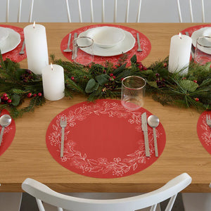 The CHILEWICH - WREATH PRINT PLACEMAT is a round, red placemat featuring a white botanical wreath with leaves, berries, and pine branches around the edge. Its center is plain red for a festive, elegant look.
