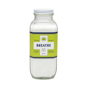 A shelf displays BAUDELAIRE Breathe Bath Seltzer alongside essential oils, a quirky face-shaped pot with a plant, and a wooden brush. The background showcases a geometric pattern.