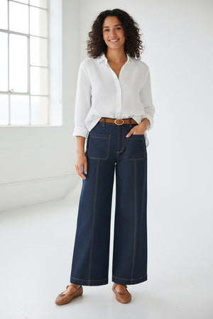 A woman with curly hair stands smiling in a bright room, wearing the MERSEA Andie Woven Pant in Rinse—high-waisted, wide-leg trousers with front pockets—paired with a white button-up shirt tucked in, a brown belt, and brown woven flats. One hand rests in her pocket.