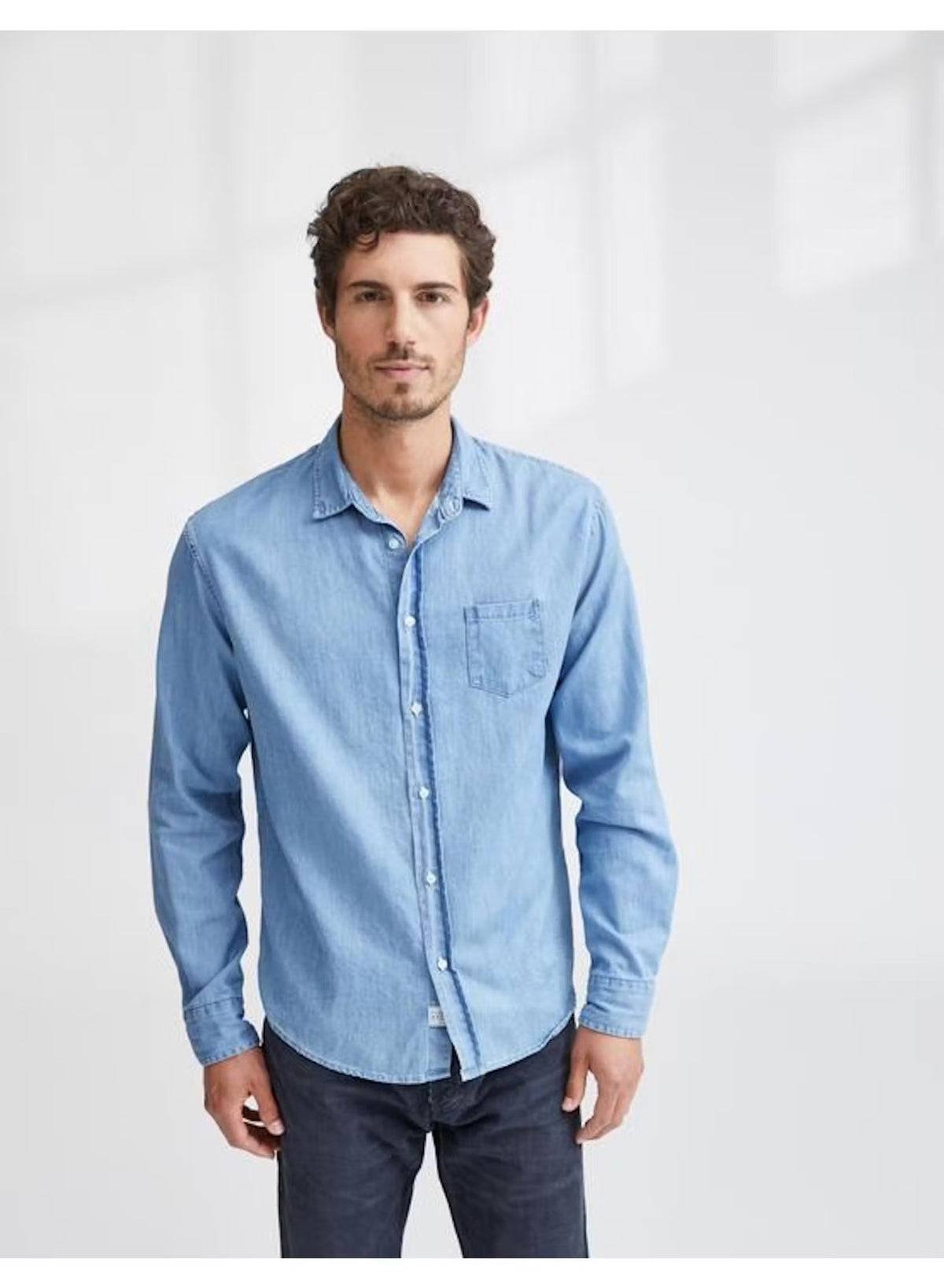 A man with short curly hair wears the FRANK & EILEEN LUKE Men’s Classix Button Up Shirt in Famous Denim Classic Blue Wash, paired with dark pants. He stands against a plain white background, facing forward with a neutral expression. This classic button-up from FRANK & EILEEN offers versatile, effortless style perfect for everyday wear.