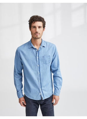 A man with short curly hair wears the FRANK & EILEEN LUKE Men’s Classix Button Up Shirt in Famous Denim Classic Blue Wash, paired with dark pants. He stands against a plain white background, facing forward with a neutral expression. This classic button-up from FRANK & EILEEN offers versatile, effortless style perfect for everyday wear.