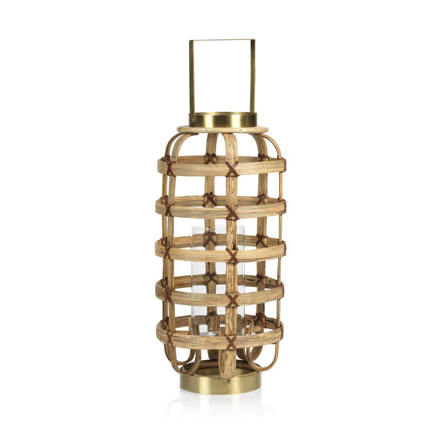 The Zodax Bamboo Lantern with Brass Trim is a decorative piece featuring a cylindrical glass holder encased in a crisscross bamboo lattice. Measuring 24.13 cm x 24.13 cm x 52.07 cm, it boasts metallic top and base elements with a convenient handle for easy carrying, seamlessly blending rustic and modern design elements.