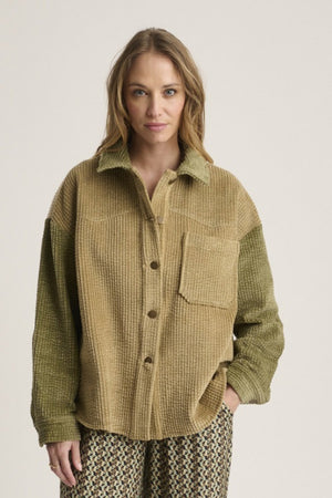 A woman with light brown hair wears the STORIATIPIC Corduroy Multi Shacket by STORIATIPIC—a tan and olive green button-up jacket crafted from soft cotton corduroy with a single chest pocket—paired with patterned pants. She stands against a plain light background, gazing at the camera with a neutral expression.