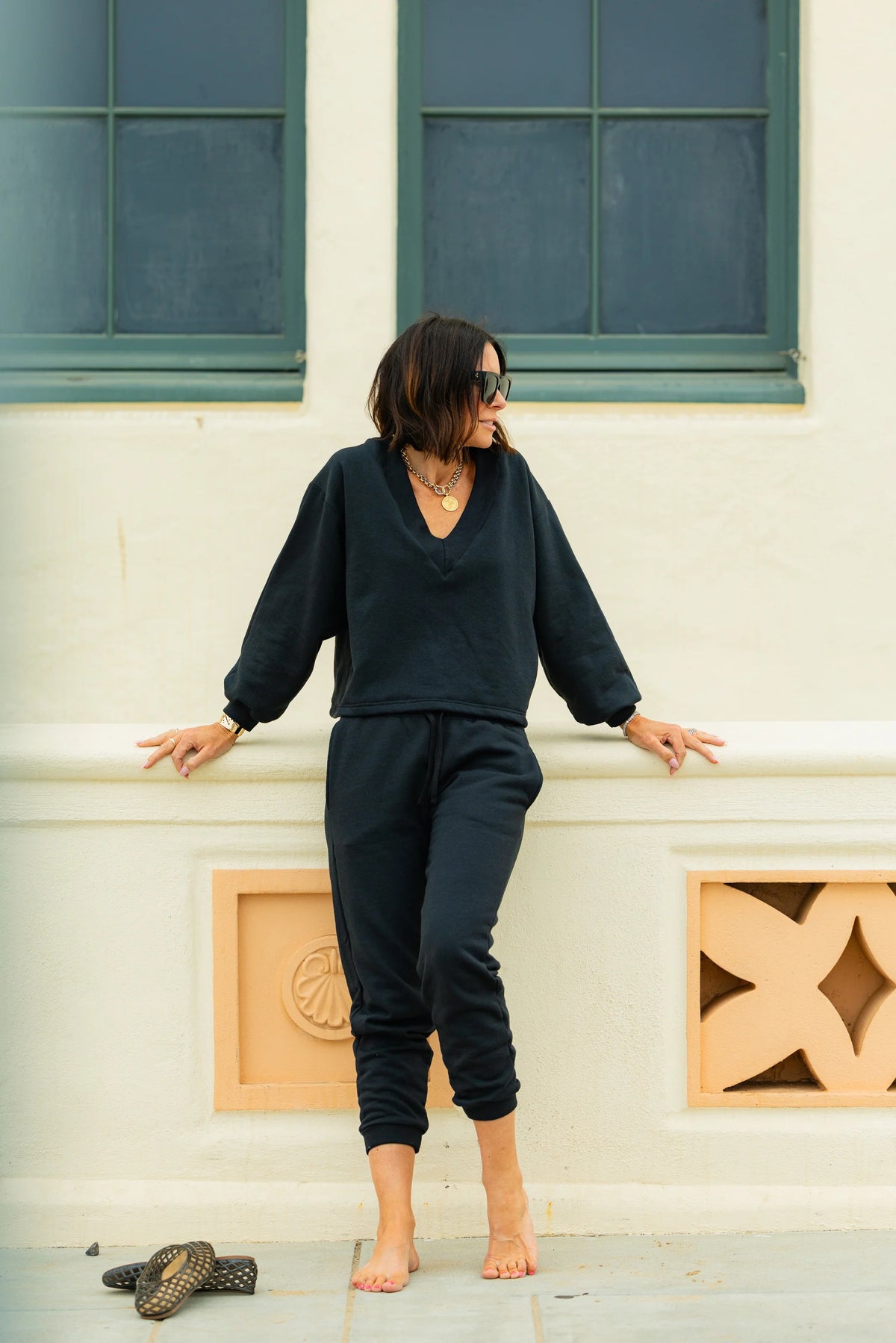 A woman stands indoors by a large window, wearing the KMJ - TUCKER V NECK SWEATSHIRT IN BLACK from KMJ, paired with blue jeans and black ankle boots. With her hands in her pockets and a soft smile on her face, she is bathed in sunlight streaming into the modern, minimalist room.