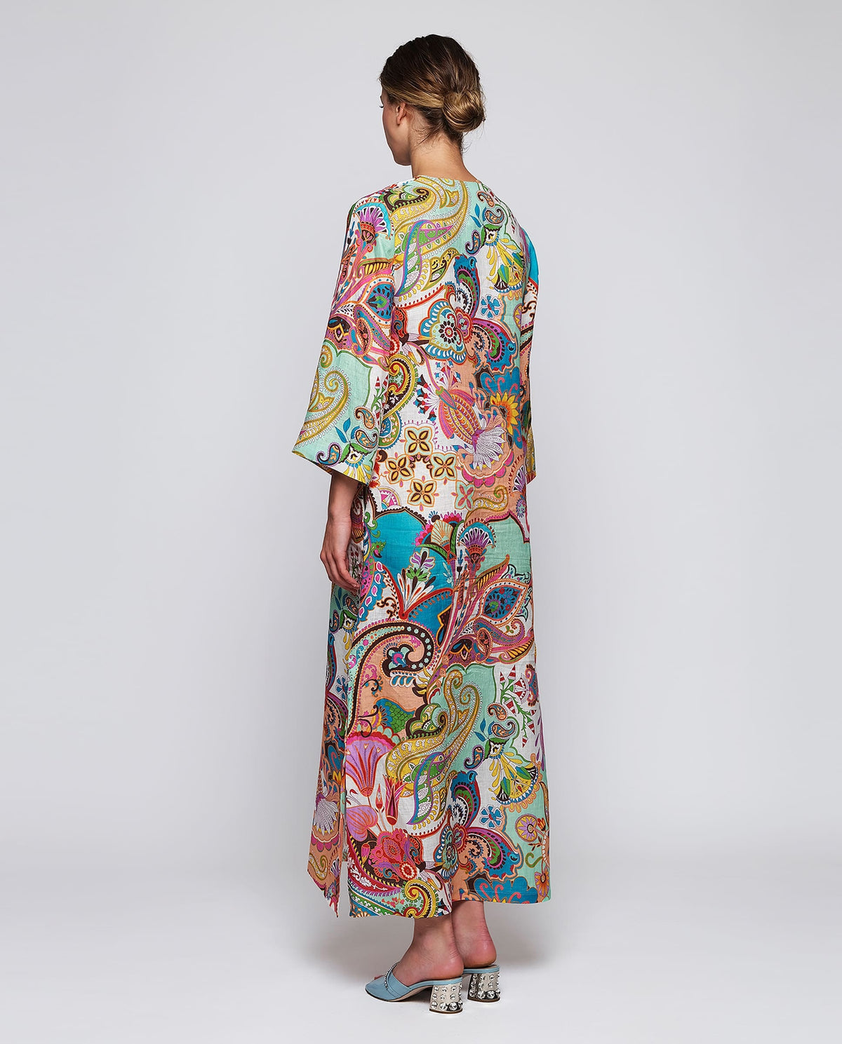 A woman stands facing forward wearing the MIRTO 1956 Multi Color Paisley Kaftan Style Dress with wide sleeves and a V-neckline, paired with blue heeled sandals, against a plain white background.