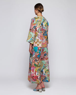 A woman stands facing forward wearing the MIRTO 1956 Multi Color Paisley Kaftan Style Dress with wide sleeves and a V-neckline, paired with blue heeled sandals, against a plain white background.