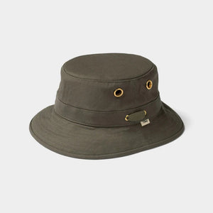 A person wearing the TILLEY - ICONIC T1 BUCKET HAT IN OLIVE with yellow eyelets and a white crew-neck shirt looks slightly to the side against a plain light background. The TILLEY hat offers UPF 50+ sun protection.