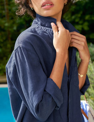 A woman in the FRANK & EILEEN Eileen Relaxed Button Up Shirt in Linen from the Tuscany Getaway Set stands poolside among greenery, wearing wide-leg pants, sandals, sunglasses, and sporting short curly hair as she gazes upward.