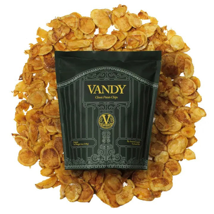 A black bag labeled &quot;ANCIENT CRUNCH - VANDY TRADITIONAL POTATO CHIPS&quot; stands upright with a stylish design, surrounded by a scattered pile of golden-brown potato chips in front of it.