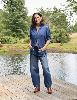 A close-up of a person wearing the FRANK & EILEEN Brennan Everyday Barrel Jean in California Vintage Denim 1993 Wash, paired with a loose blue shirt and one arm resting by their side.