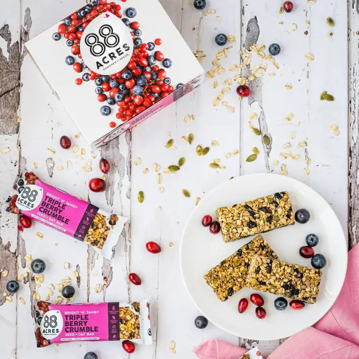 An 88 ACRES - TRIPPLE BERRY CRUMBLE Seed + Oat Bar rests on a light wooden surface, with its package partially open to show a portion of the nut-free and vegan granola bar inside. The packaging is white, featuring a vibrant purple section that displays the product name and various icons.