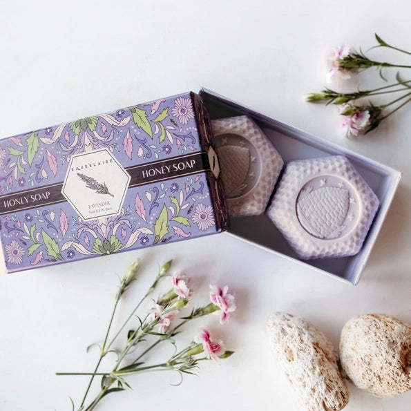 The BAUDELAIRE - LAVENDER HONEY SOAP 3.5 OZ, 2 BAR GIFT BOX features two hexagonal, triple-milled soaps with a beehive design, nestled in a decorative purple floral box and partially opened to reveal the lavender-colored bars inside.