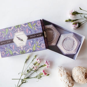The BAUDELAIRE - LAVENDER HONEY SOAP 3.5 OZ, 2 BAR GIFT BOX features two hexagonal, triple-milled soaps with a beehive design, nestled in a decorative purple floral box and partially opened to reveal the lavender-colored bars inside.