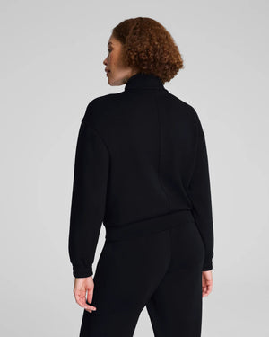 A person with curly hair models the SPANX AIRESSENTIALS BRUSHED FULL ZIP JACKET in black with matching pants, standing against a light gray background—highlighting chic loungewear from SPANX.