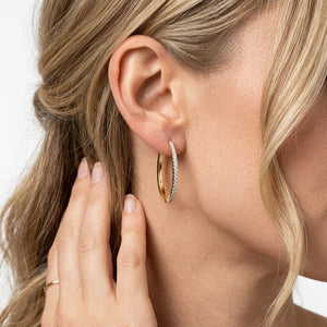 A close-up of a woman with blonde hair wearing the ELLA STEIN - SLIM PAVE DIAMOND HUGGIE (LARGE) by ELLA STEIN, a gold plated hoop earring set with small natural diamonds, as she touches her ear with her left hand.