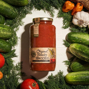 A 32oz jar of THE REAL DILL - ORIGINAL BLOODY MARY MIX by THE REAL DILL is displayed in a sunlit kitchen, surrounded by fresh tomatoes, cucumbers, dill, garlic, and a classic Bloody Mary cocktail.