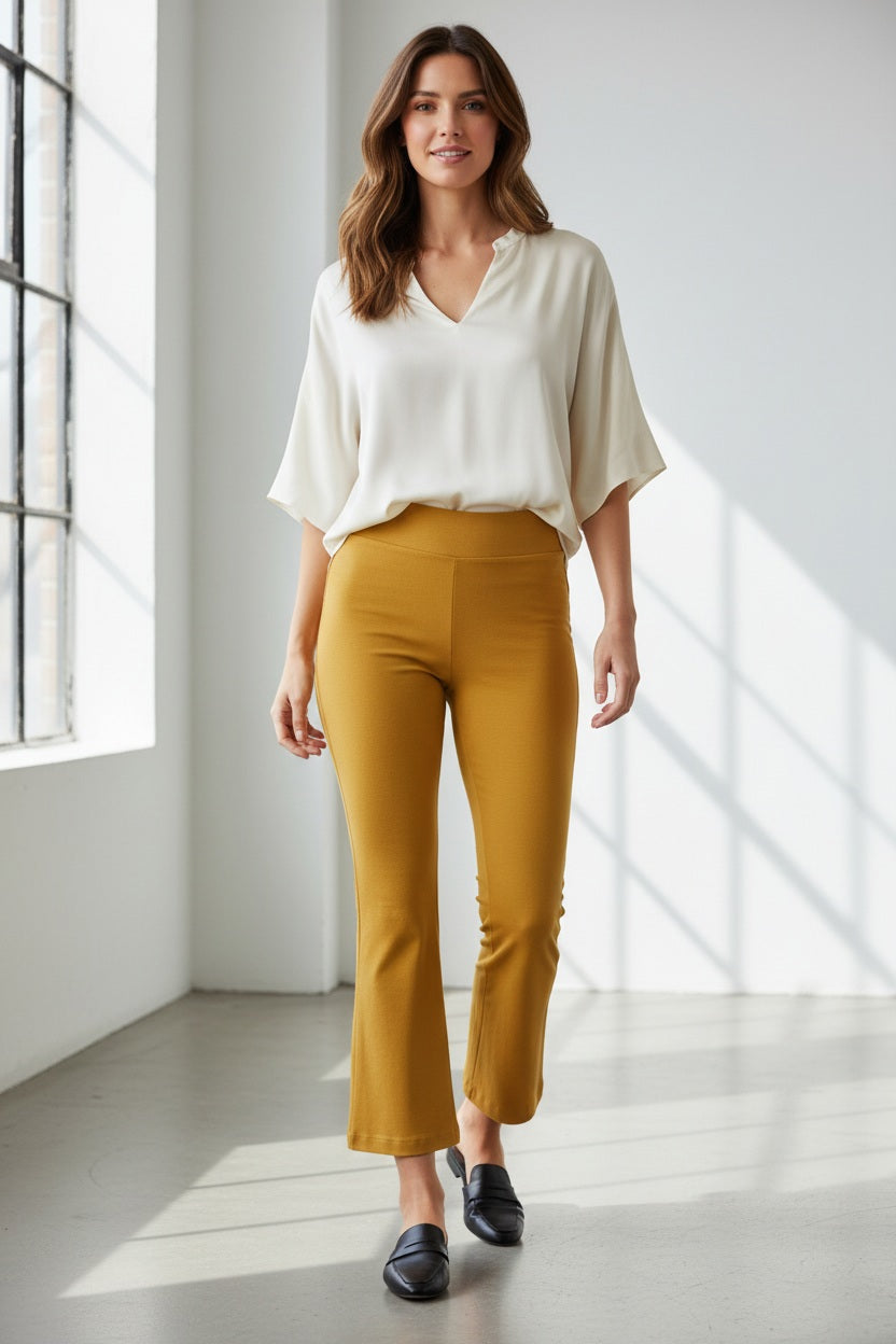 A woman stands indoors by a large window, wearing a long-sleeve cream blouse and the KRAZY LARRY, INC PULL ON WIDE ANKLE PANT in black, which features a tummy tuck design. She completes her look with black loafers and smiles slightly, her wavy brown hair catching the sunlight that casts shadows on the white floor and wall.