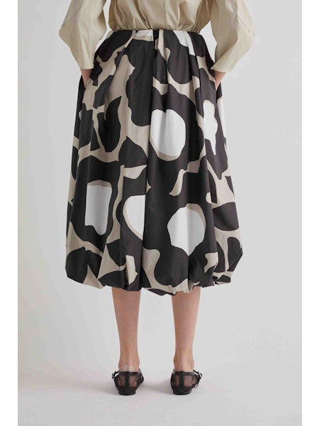A woman stands against a plain background wearing a beige long-sleeve top, black sandals, and the MEIMEIJ BLACK MOONBEAM SKIRT by MEIMEIJ—a voluminous midi skirt featuring large white and gray abstract shapes.