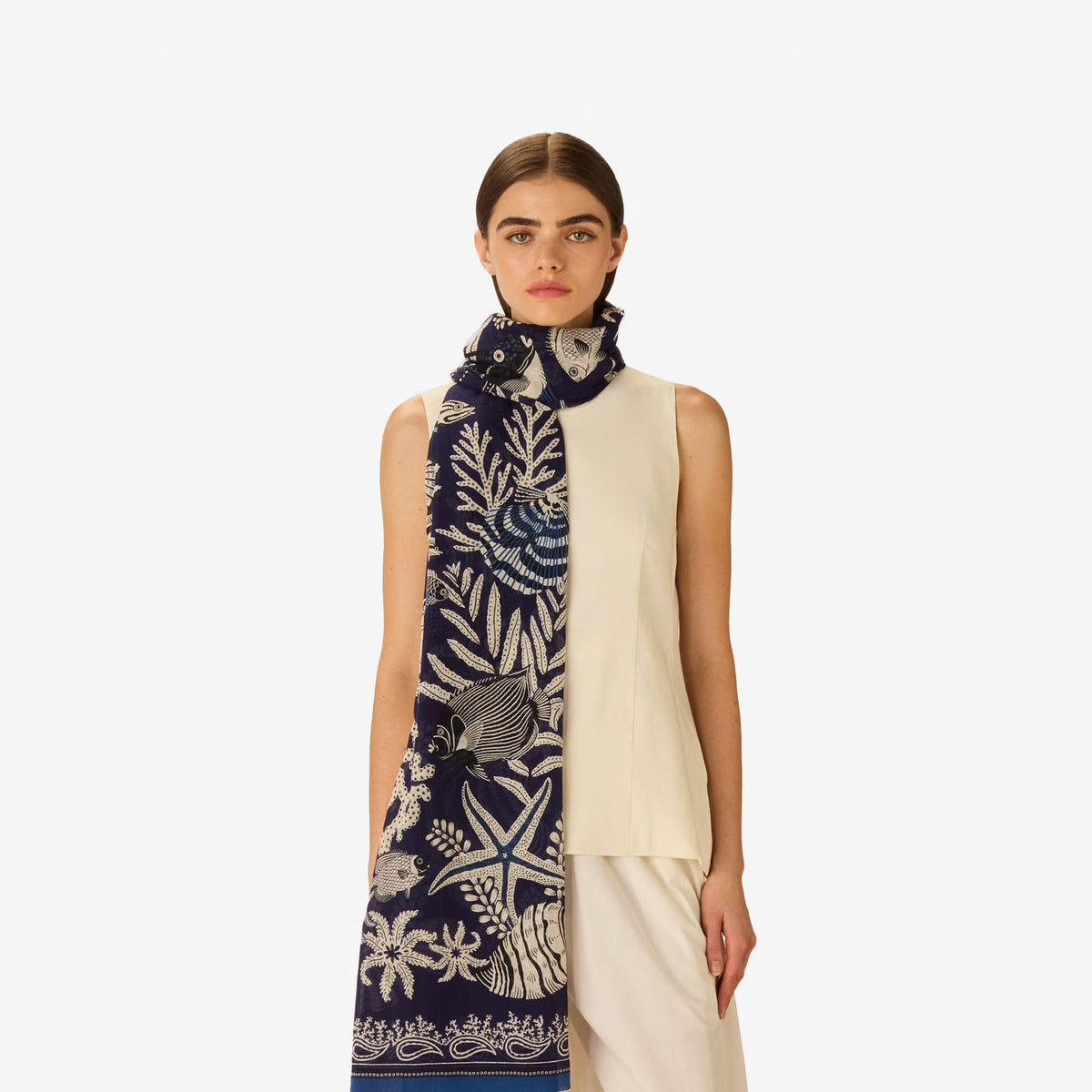 A woman faces forward, wearing a sleeveless cream and navy top with white botanical and animal motifs, accessorized with the INOUI EDITIONS - NEPTUNE SCARF IN NAVY by INOUI, draped loosely around her neck.