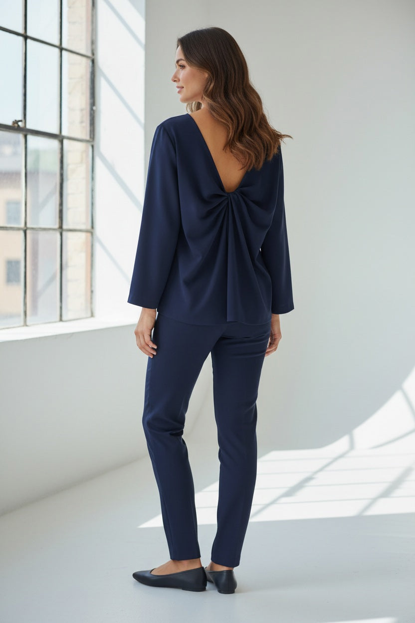 A person wears the MEIMEIJ TOP WITH KNOT BACK DETAIL in bright orange, paired with matching pants, standing against a plain light grey background. Their face is partially out of frame.