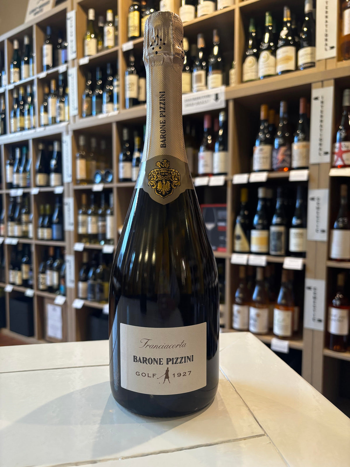 A dark bottle of BARONE PIZZINI ‘GOLF’ FRANCIACORTA from WINE: WINEBOW, featuring a gold foil top, black and gold crest near the neck, and a label reading "Franciacorta Barone Pizzini Golf 1927.
