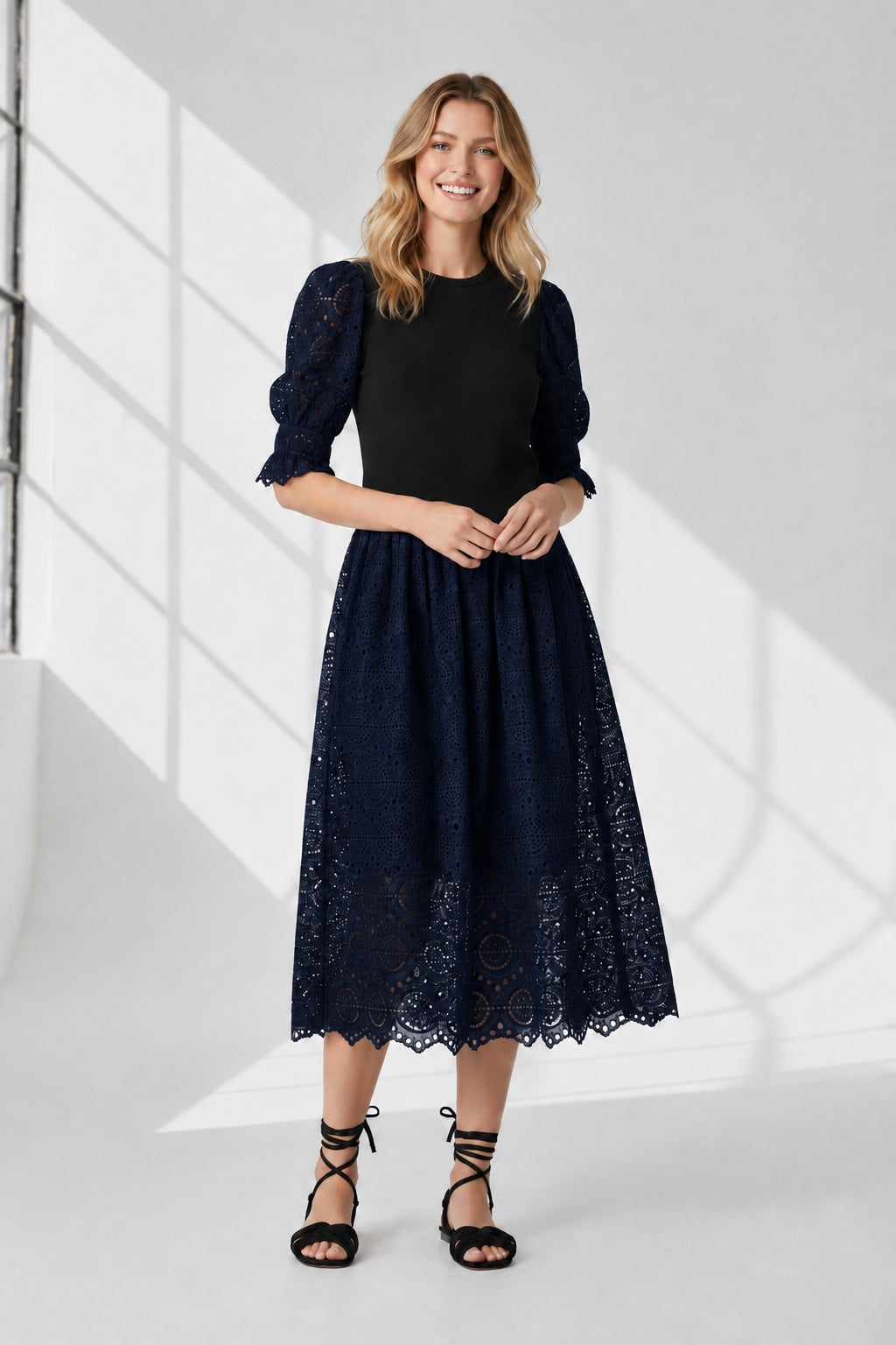 A woman stands against a white background wearing the SFIZIO - BRODERIE ANGLAISE MIDI DRESS by SFIZIO, featuring puffed sleeves and a lace skirt, paired with black strappy sandals.
