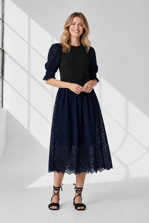 A woman stands against a white background wearing the SFIZIO - BRODERIE ANGLAISE MIDI DRESS by SFIZIO, featuring puffed sleeves and a lace skirt, paired with black strappy sandals.