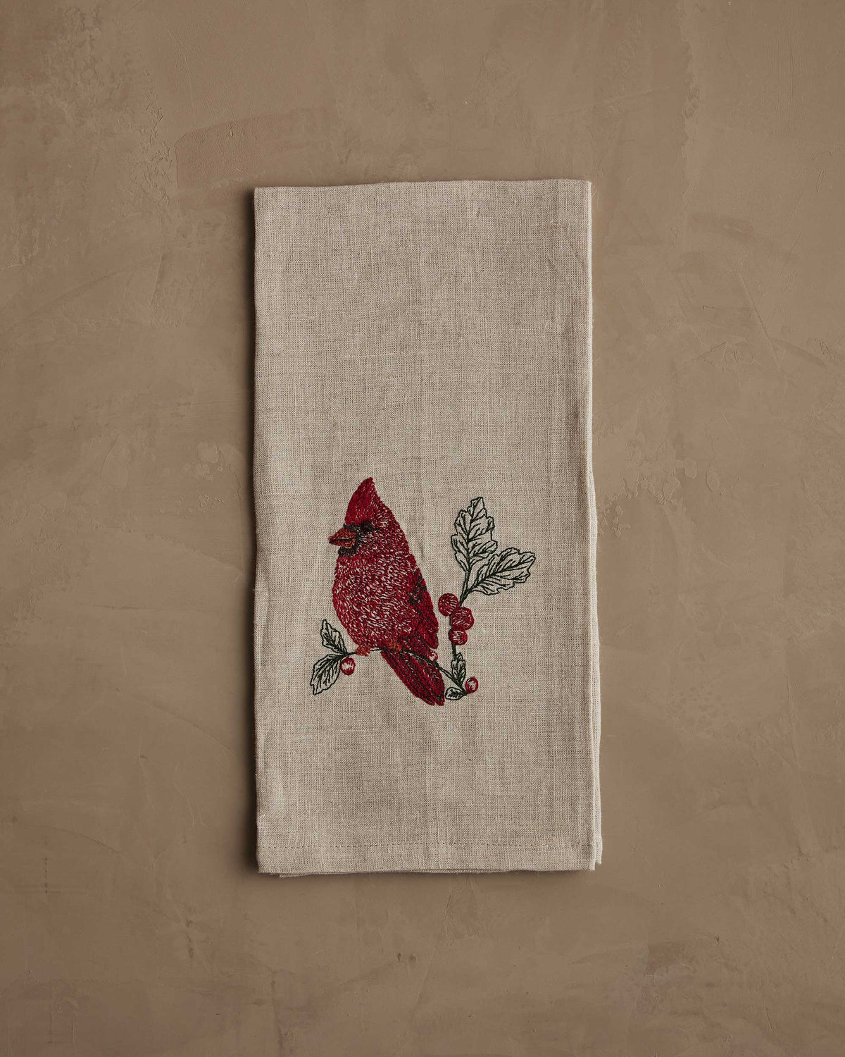 A RED CARDINAL LINEN TEA TOWEL by CORAL & TUSK made of beige natural flax fiber, embroidered with a red cardinal perched on a branch with berries and green leaves.