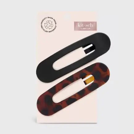 Two sustainable rectangular hair clips, one solid black and the other with a brown and black tortoiseshell pattern, rest on a light pink card. The card emphasizes the recycled materials of "KITSCH - FLAT LAY CLAW CLIP 2 PC," showcasing their style and eco-friendly commitment.