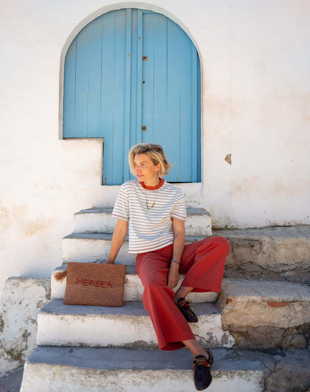 A smiling person with short blond hair wears the MERSEA Deauville Ringer Tee in Slate Blue Persimmon and red pants, holding a woven clutch while standing outside near white stairs and a blue door on a sunny day.