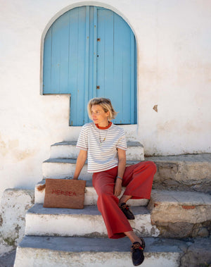 A smiling person with short blond hair wears the MERSEA Deauville Ringer Tee in Slate Blue Persimmon and red pants, holding a woven clutch while standing outside near white stairs and a blue door on a sunny day.