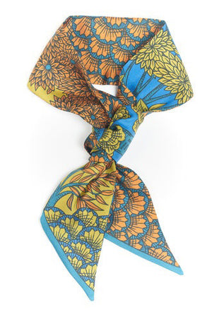 A person with dark hair styled in a low bun, adorned with the INOUI EDITIONS - MAGNUS SMALL SILK RIBBON featuring orange and green designs on a blue base, wearing a ribbed, long-sleeved maroon top. The timeless accessory from INOUI, made from 100% silk, is shown from the side against a plain white background.