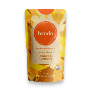 A carton of BRODO BROTH- DEEPLY ROOTED TURMERIC & GINGER BONE BROTH ORGANIC by BRODO BROTH COMPANY rests on a blue tray, surrounded by fresh turmeric and ginger roots—an ideal choice for those seeking a keto-friendly option.