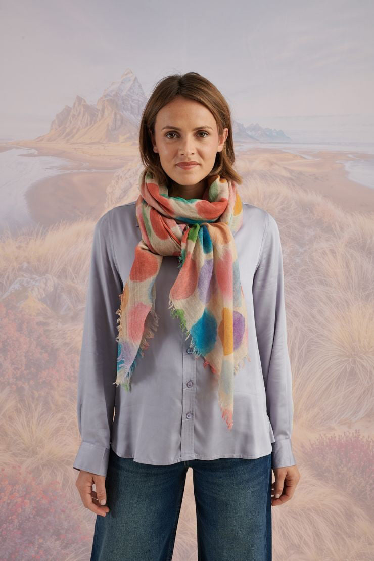 A woman with blonde hair displays the STORIATIPIC - BETTY WOOL SCARF, a large rectangular wool accessory with earth-toned polka dots, set against a scenic mountain landscape.
