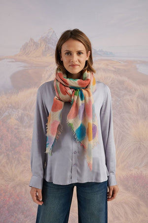 A woman with blonde hair displays the STORIATIPIC - BETTY WOOL SCARF, a large rectangular wool accessory with earth-toned polka dots, set against a scenic mountain landscape.