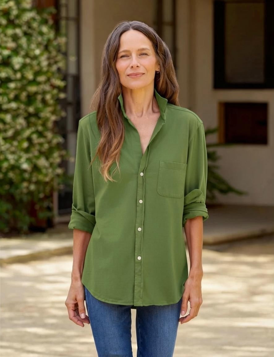 A woman with long brown hair stands outdoors, smiling slightly at the camera while wearing the FRANK & EILEEN Waverly Button Up Tee in Vintage Jersey Nori and blue jeans. In the background, there are plants and a building.