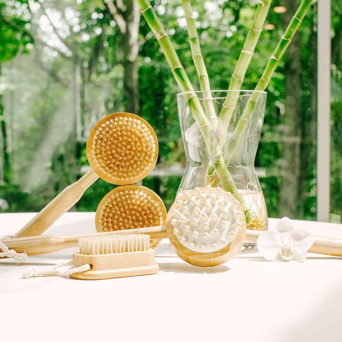 A woman with a towel on her head relaxes in a bathtub, gently scrubbing her hand with the BAUDELAIRE - BAMBOO 4” NAIL BRUSH from BAUDELAIRE. She is wearing a white floral top and is surrounded by neutral-colored tiles.