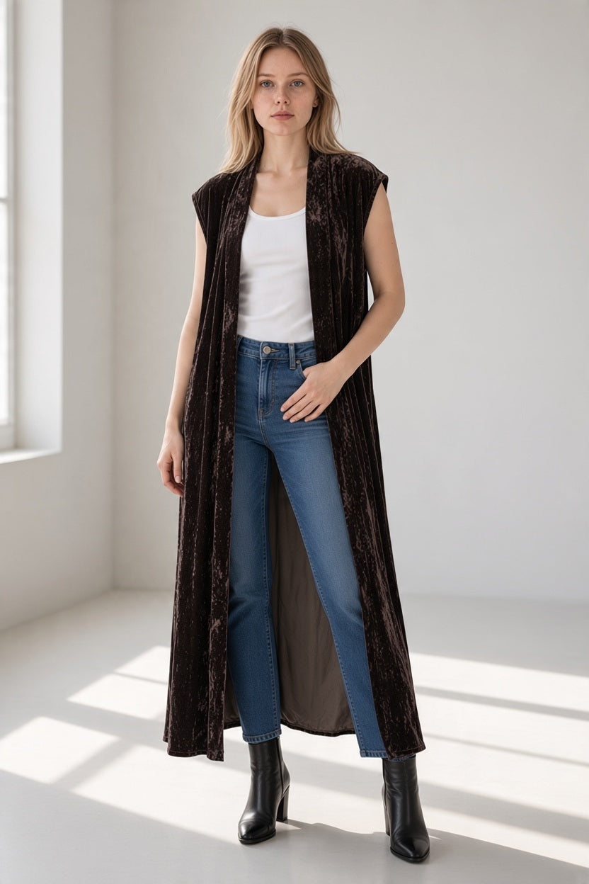 A woman wears an elegant, dark brown GAYNOR BONGARD POETA VELVET GILET IN BROWNIE over a white tank top and blue jeans with black heeled boots.