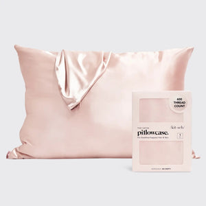 A silky KITSCH satin pillowcase in light pink is elegantly displayed against a plain backdrop, symbolizing luxury. A matching KITSCH frizz-free satin scrunchie is draped over it, while a box labeled "600 Thread Count" sits beside the pillow containing another beautiful satin pillowcase.