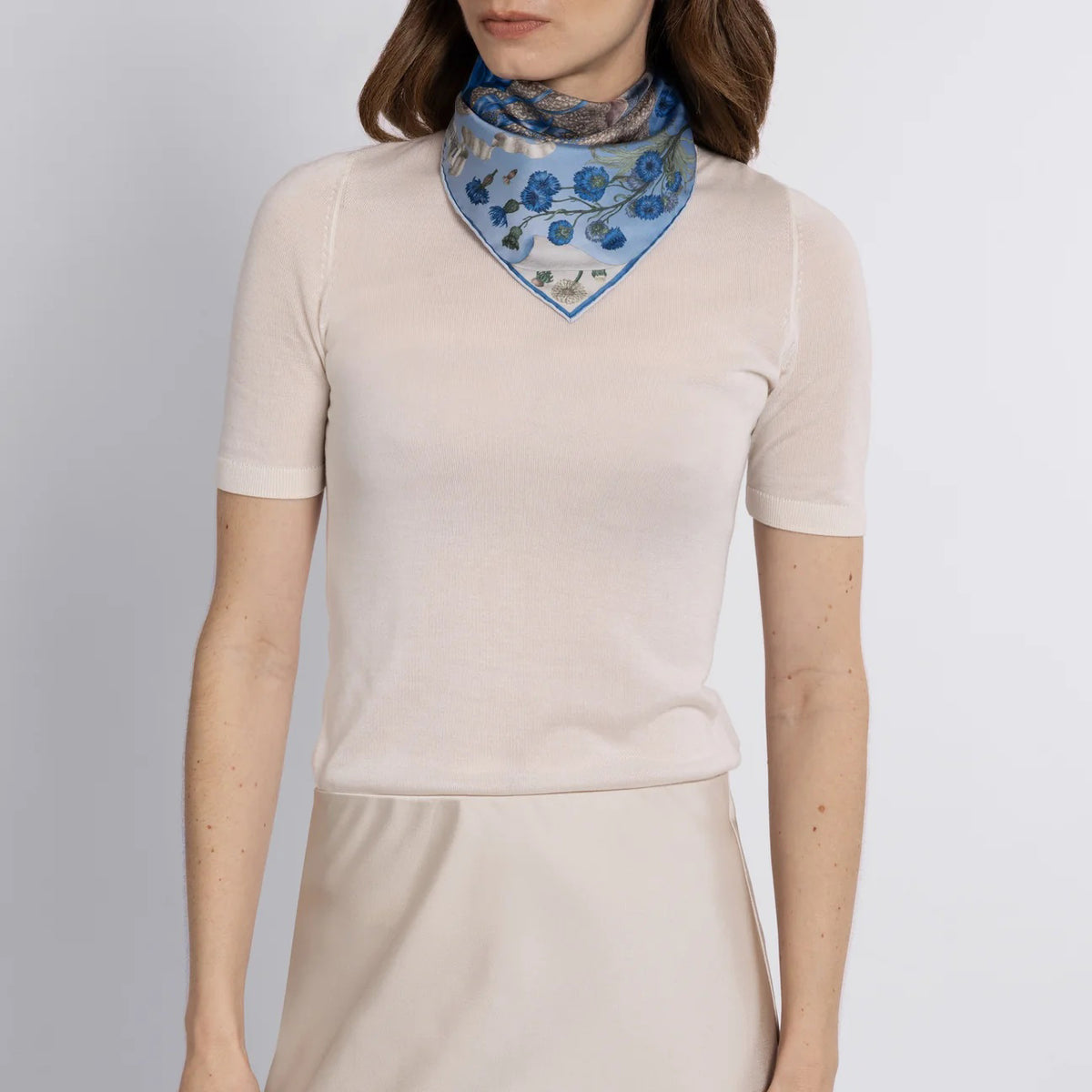 The SABINA SAVAGE - LONDON BRIDGE IS FALLING DOWN SILK TWILL SCARF 42CM POWDER/FROST from SABINA SAVAGE ENGLAND is artfully folded into a floral shape, showcasing a blue and white pattern with flowers, baby lambs, and intricate details bordered by a bold blue.