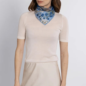 The SABINA SAVAGE - LONDON BRIDGE IS FALLING DOWN SILK TWILL SCARF 42CM POWDER/FROST from SABINA SAVAGE ENGLAND is artfully folded into a floral shape, showcasing a blue and white pattern with flowers, baby lambs, and intricate details bordered by a bold blue.