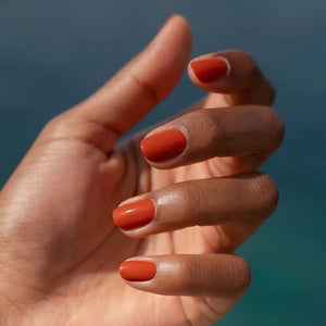 A hand with a glossy coat of MANUCURIST - GREEN™ NAIL POLISH IN TERRACOTTA by MANUCURIST INC touches an ear adorned with a chunky, abstract silver earring against brown skin.