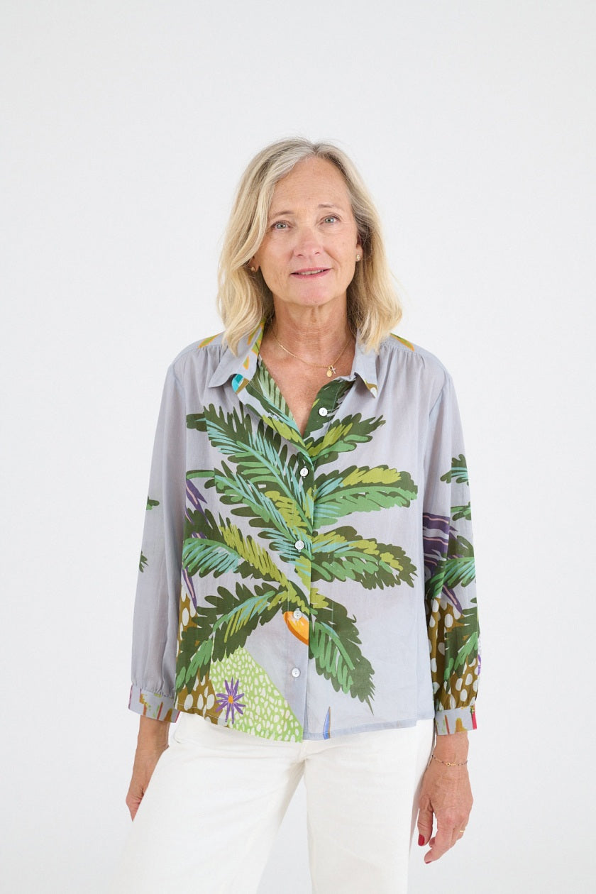A woman with shoulder-length blonde hair wears the STORIATIPIC - SUZY CALI BLOUSE by STORIATIPIC, a light gray cotton top with a colorful palm tree print, paired with white pants and smiling gently against a plain white background.