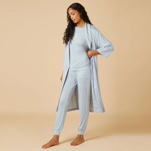Wearing light blue and white striped pajamas, a person stands barefoot against a beige background, draped in the SOFTIES Dream Shawl Collar Robe in Sky Blue Stripe. This lightweight robe from SOFTIES adds an extra layer of silky smooth comfort to the cozy look.