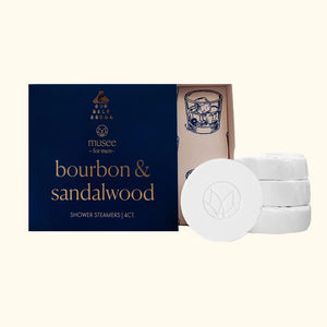 A blue box labeled “MUSEE - BOURBON & SANDALWOOD SHOWER STEAMERS” by MUSEE sits beside four white round steamers and an open lid adorned with a drawing of a whiskey glass, offering an indulgent aromatherapy shower experience enhanced with sandalwood essential oil.
