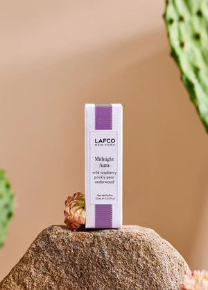 A person holds a 10ml bottle of LAFCO Fine Fragrance, Midnight Aura, on their bare shoulder against a soft purple background.