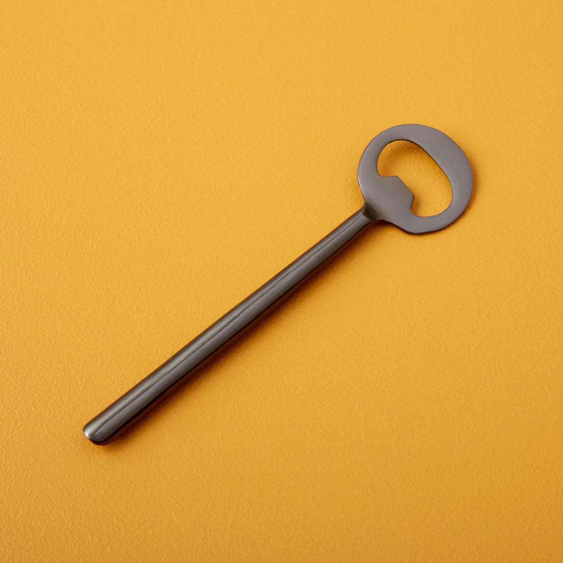 The BE HOME, INC MATTE STAINLESS BOTTLE OPENER with a sleek circular head and elongated handle rests on a vibrant mustard-yellow surface. This minimalistic silver-colored piece is a standout addition to any barware collection.